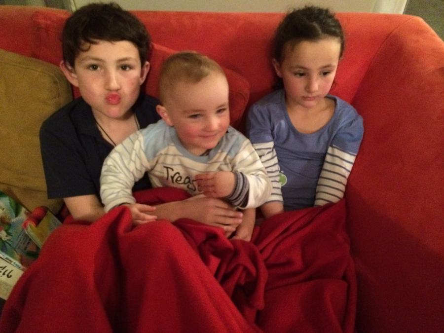 Three children under a red blanket on the sofa